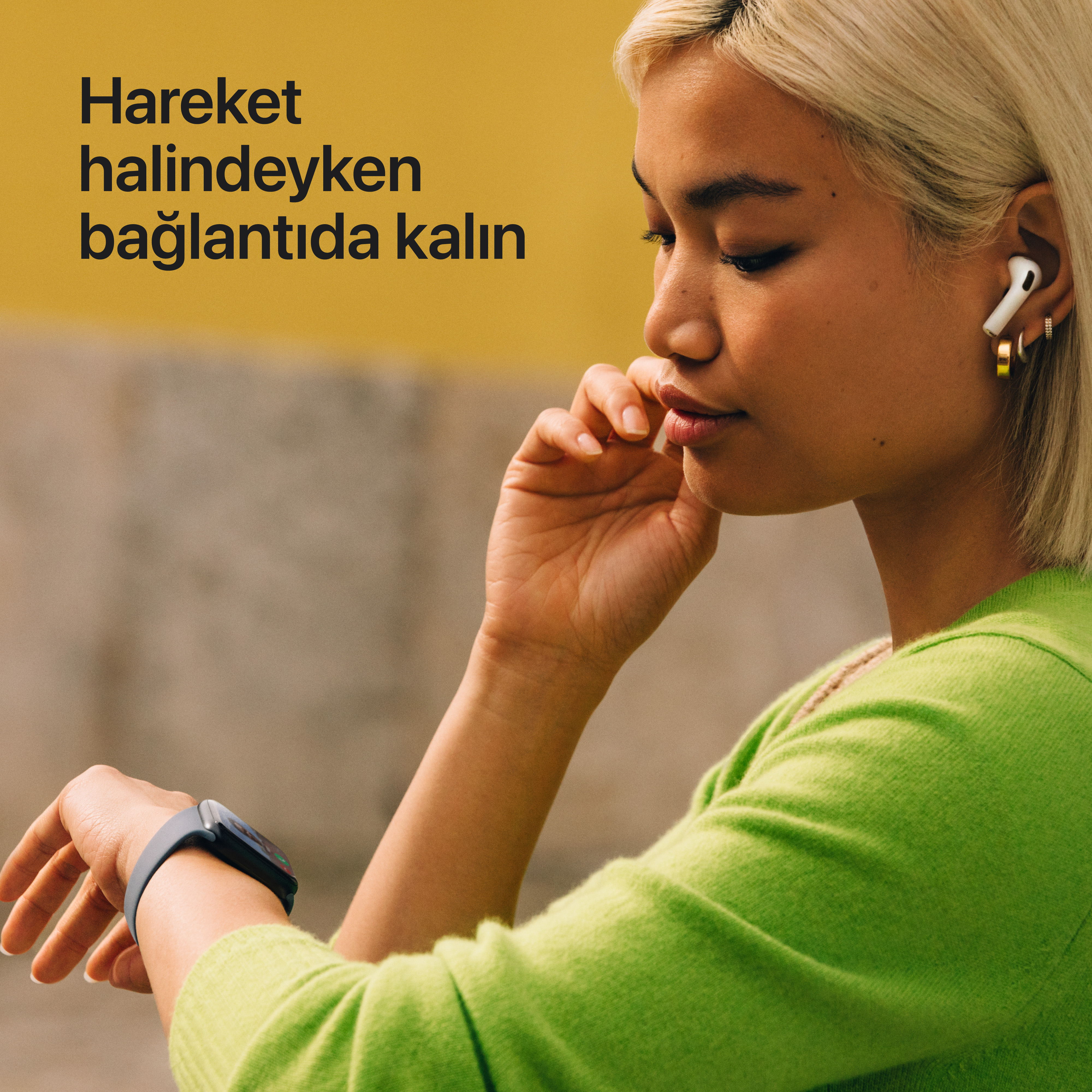 Airpods ve akıllı saat takan kadın, yüzüne dokunuyor. Yeşil gömlek. Metinli sarı arka plan.