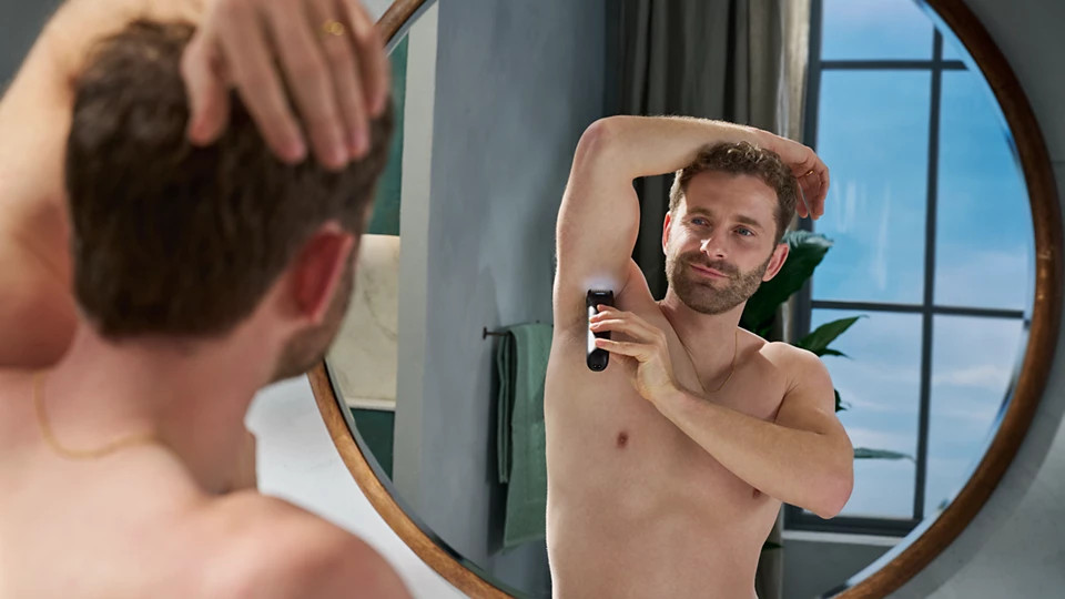 Mężczyzna goli pachy trymerem przed lustrem. Lustro odbija jego plecy. W tle okno.