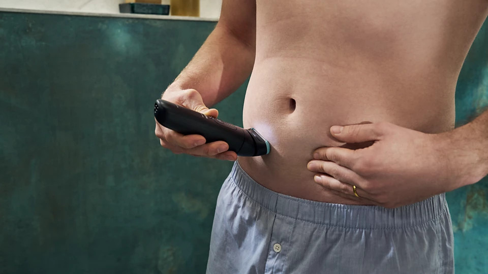 Mężczyzna goli brzuch elektryczną maszynką do strzyżenia. Ma na sobie jasnoniebieskie spodnie od piżamy.