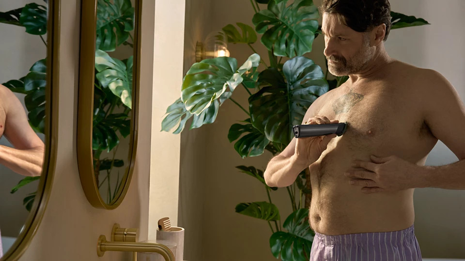 Mężczyzna goli klatkę piersiową w łazience. Trzyma trymer, patrząc w lustro. Widoczne są zielone rośliny i złote elementy.