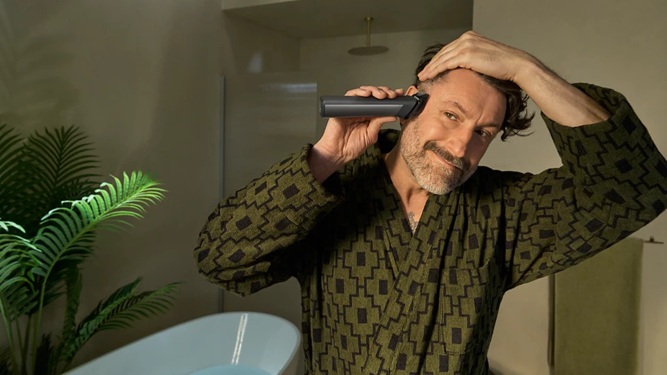 Mężczyzna w szlafroku w wzory goli włosy trymerem w łazience. Widoczna jest również roślina i wanna.