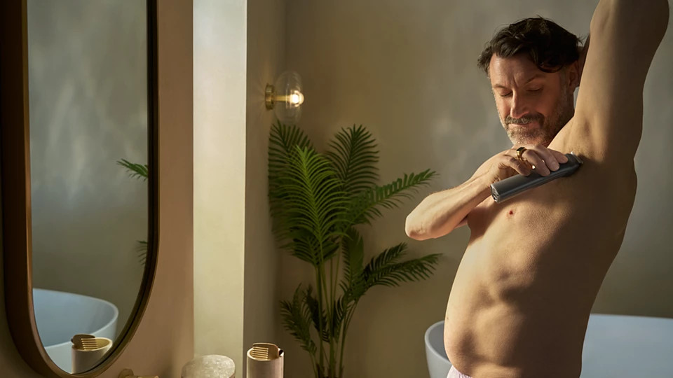 Mężczyzna goli pachy maszynką elektryczną w łazience. Widoczne lustro, roślina i lampa.