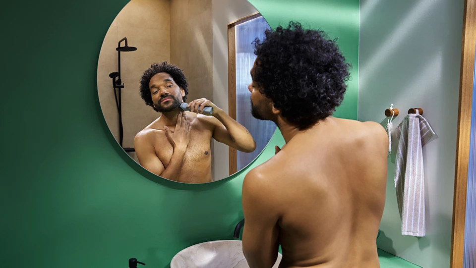 Mężczyzna goli się przed lustrem. Trzyma golarkę elektryczną przy szyi. Zielona ściana, prysznic i ręcznik widoczne w tle.
