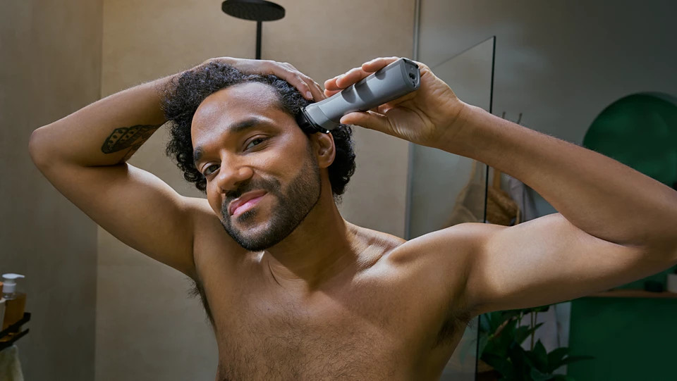 Mężczyzna goli głowę w łazience. Trzyma trymer przy głowie, uśmiechając się. Tatuaż na ramieniu. Beżowa ściana i lustro.