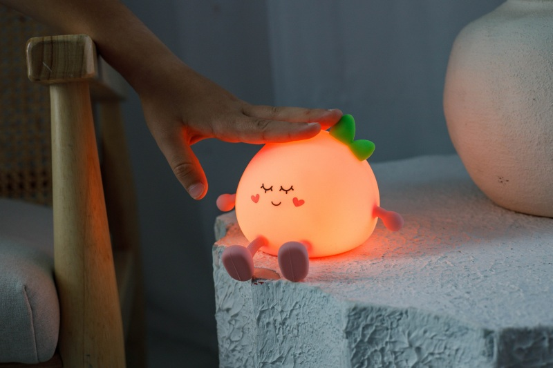 Ręka dotyka świecącej lampy w kształcie owocu. Lampa ma twarz i stopy. Drewniane krzesło i biały wazon są w tle.