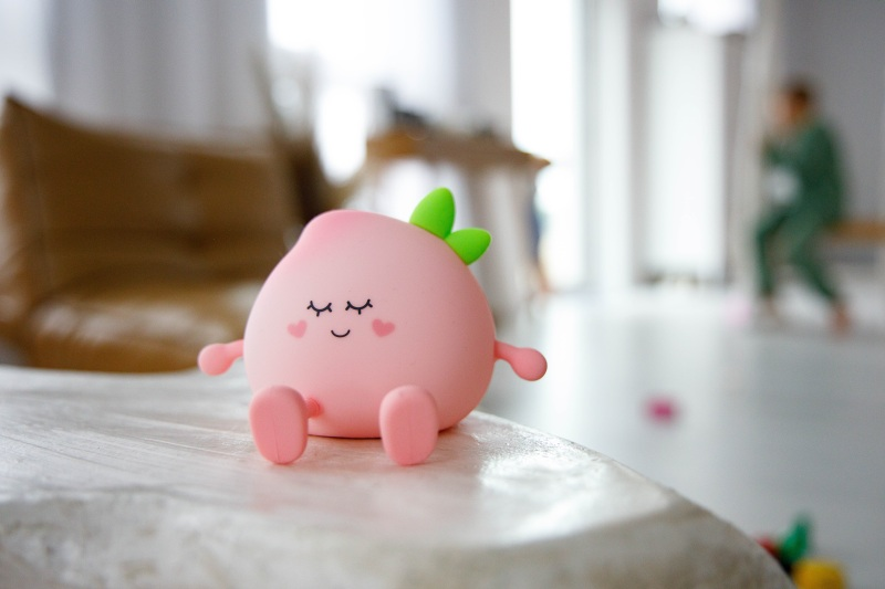 Urocza zabawka w kształcie brzoskwini z zamkniętymi oczami, małymi ramionami i nogami siedzi na stole. W tle niewyraźna osoba w zieleni.