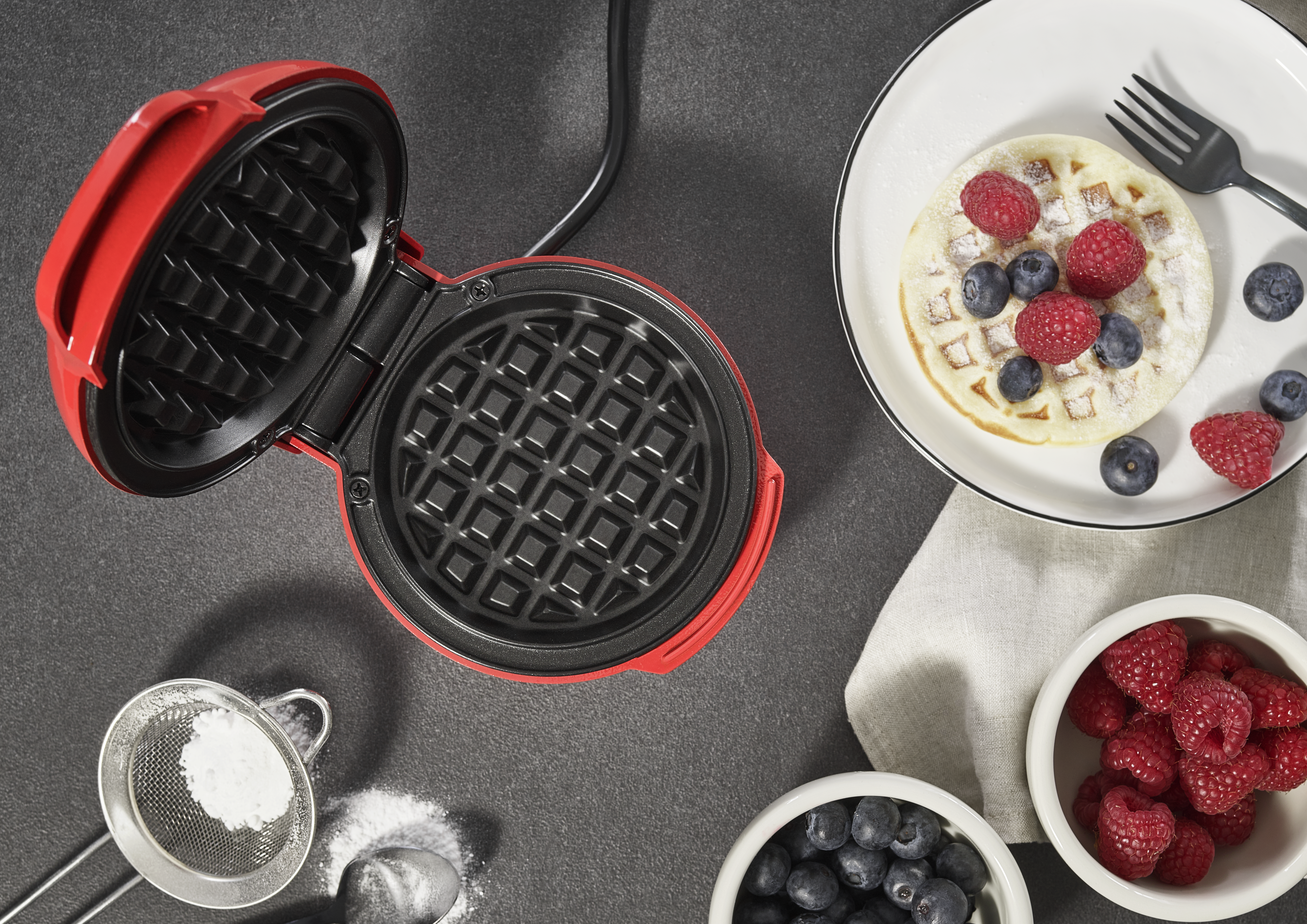 Roter Waffeleisen, Waffel auf Teller mit Beeren, Schalen mit Beeren, Puderzucker und eine Gabel auf dunkler Oberfläche.