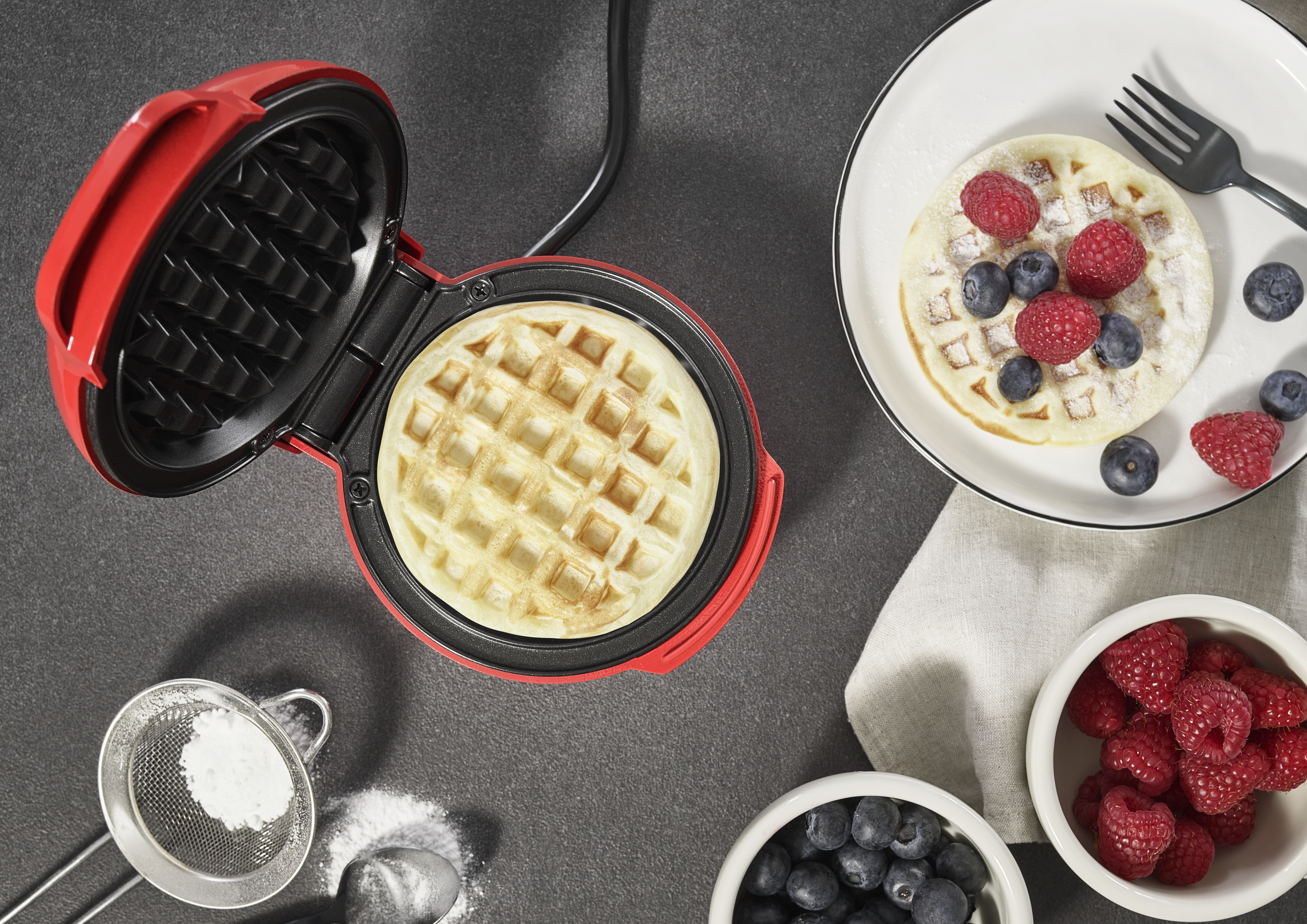 Ein Waffeleisen mit einer fertigen Waffel. Ein Teller mit Waffel, Himbeeren und Blaubeeren. Schalen mit Beeren und Puderzucker.