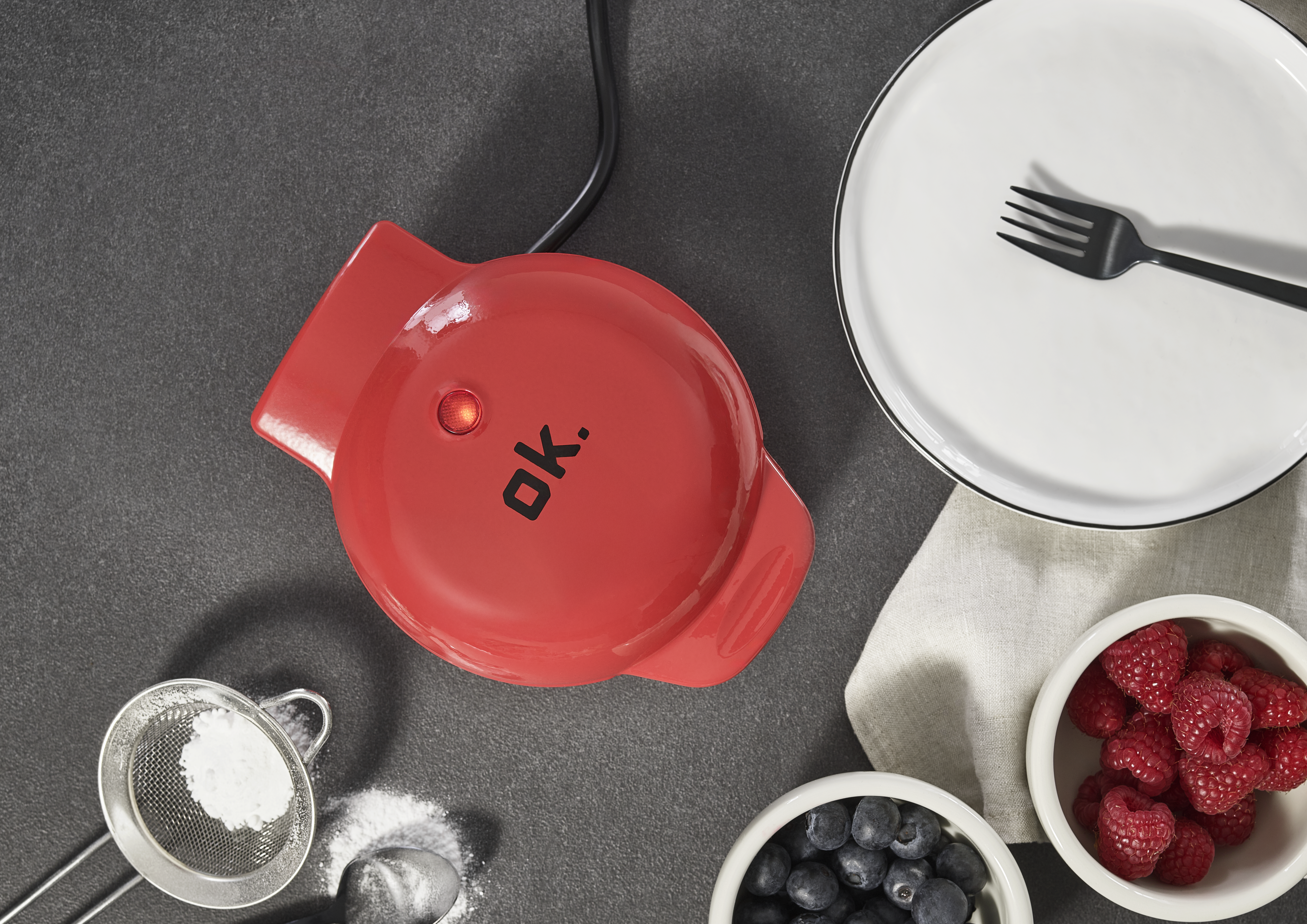 Ein roter Waffeleisen mit der Aufschrift 'ok.', ein weißer Teller mit einer Gabel und kleine Schalen mit Beeren auf einer grauen Oberfläche.