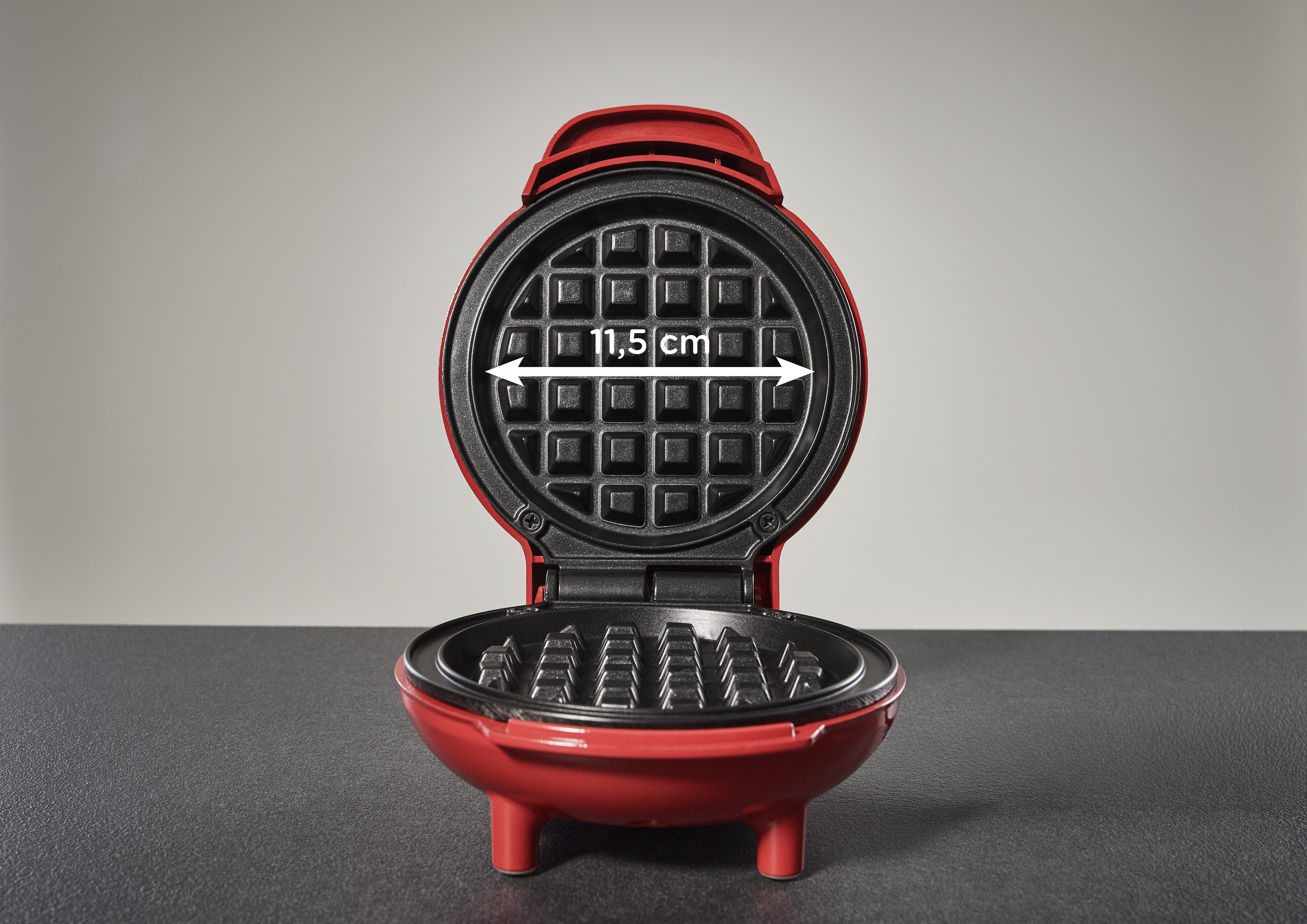 Offener roter Waffeleisen, das Waffelgitter im Inneren zeigend. Ein weißer Pfeil zeigt ein Maß von 11,5 cm an. Grauer Hintergrund, dunkle graue Oberfläche.