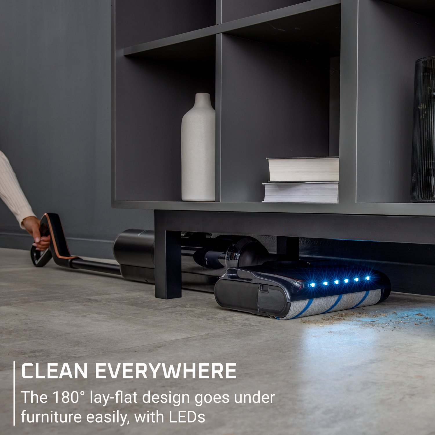 A vacuum cleaner with LED lights is under furniture. A person holds the handle. Shelves and decorative objects are in the background.