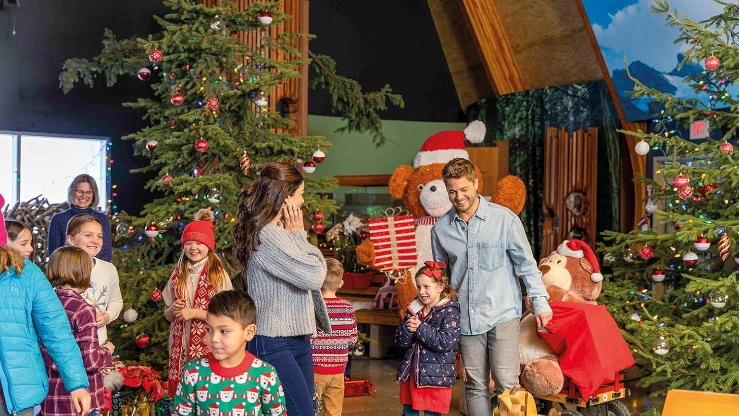 Eine Familie versammelt sich um geschmückte Weihnachtsbäume und einen großen Teddybären mit Weihnachtsmütze. Kinder tragen Winterkleidung.