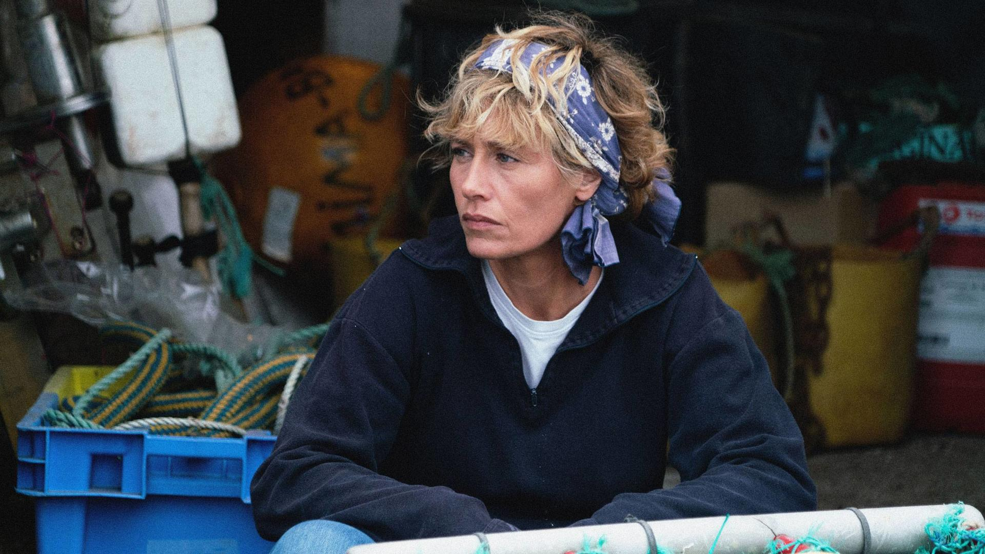 Frau mit blauem Bandana, blickt weg. Trägt einen marineblauen Pullover und ein weißes T-Shirt. Seile und Ausrüstung hinter ihr.