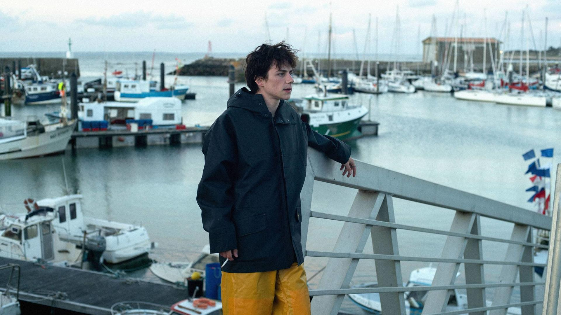 Ein junger Mann in dunkler Jacke und gelber Hose steht an einem Geländer und blickt auf Boote in einem Hafen.