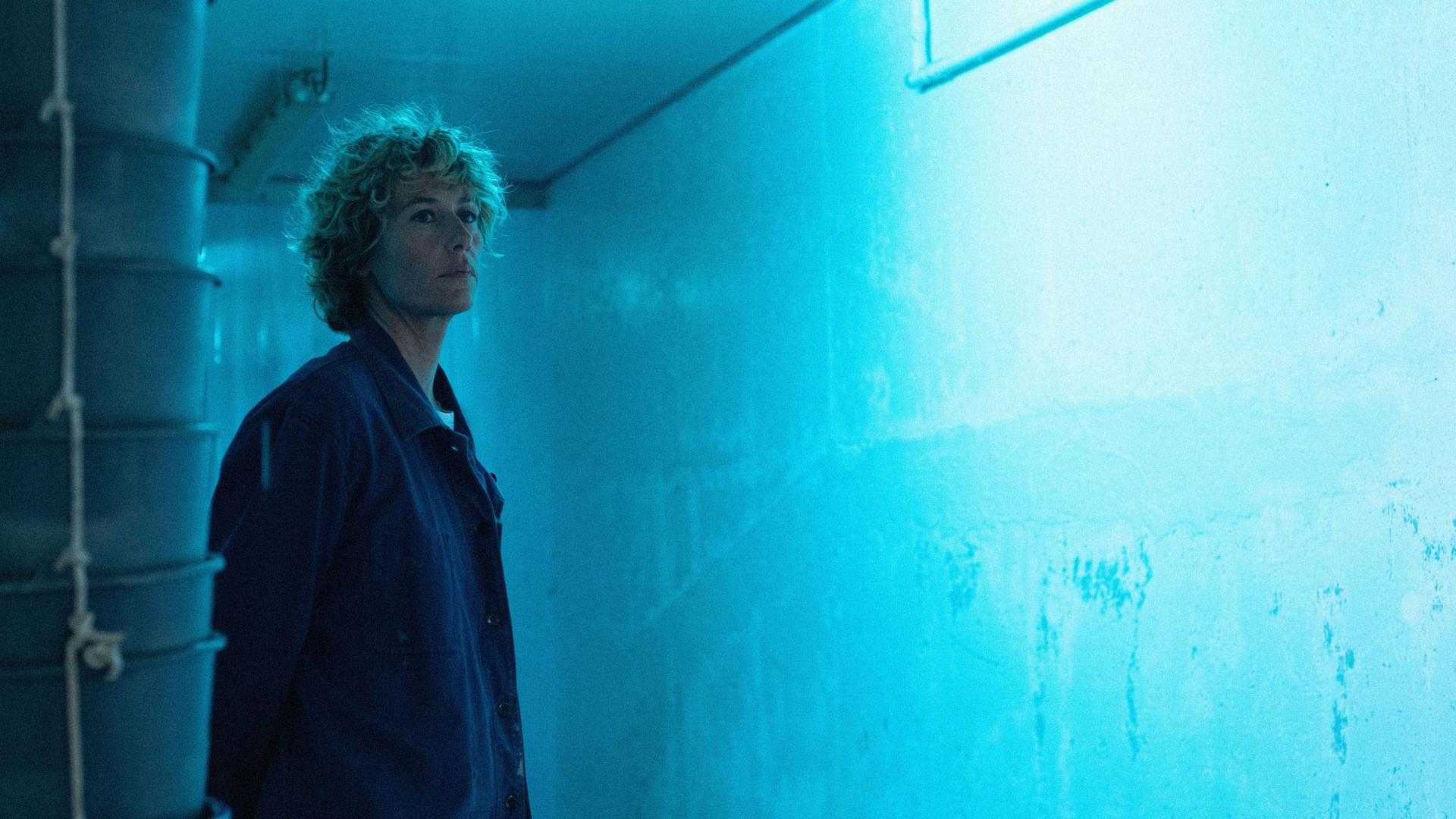 Eine Frau in einer blauen Jacke steht in einem blau beleuchteten Raum. Lockiges Haar, nach oben blickend. Hintergrund sind Eimer und eine Wand.