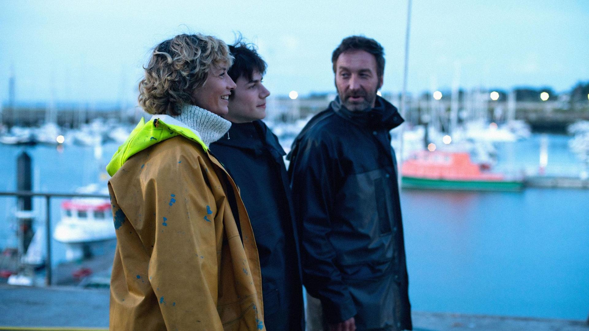 Drei Personen stehen auf einem Steg und blicken aufs Wasser. Eine Frau in gelbem Regenmantel und ein Mann in schwarzem Mantel. Boote im Hintergrund.