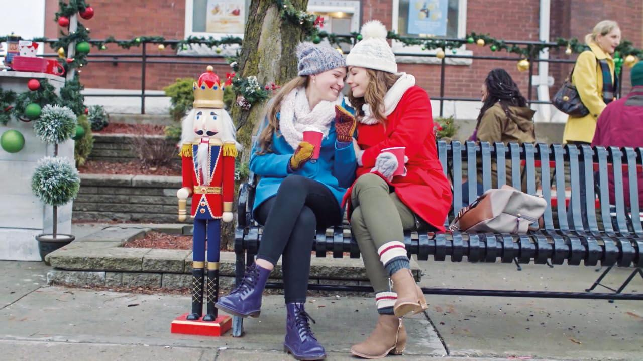 Zwei Frauen sitzen auf einer Bank und berühren sich mit den Stirnen. Ein Nussknacker steht in der Nähe. Im Hintergrund Weihnachtsdekorationen.