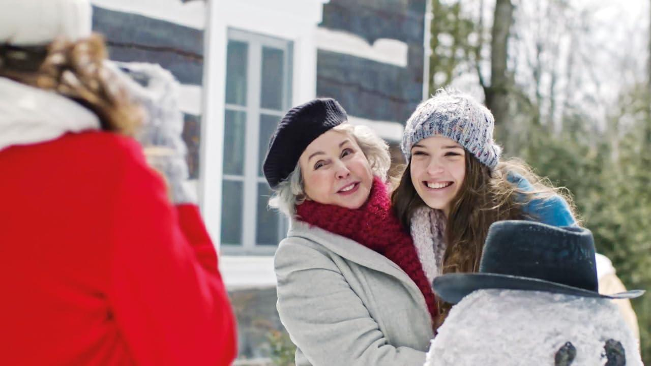 Zwei Frauen und ein Schneemann im Schnee. Eine Frau in Rot macht ein Foto von den anderen beiden, die lächeln, mit schneebedeckten Bäumen hinter ihnen.
