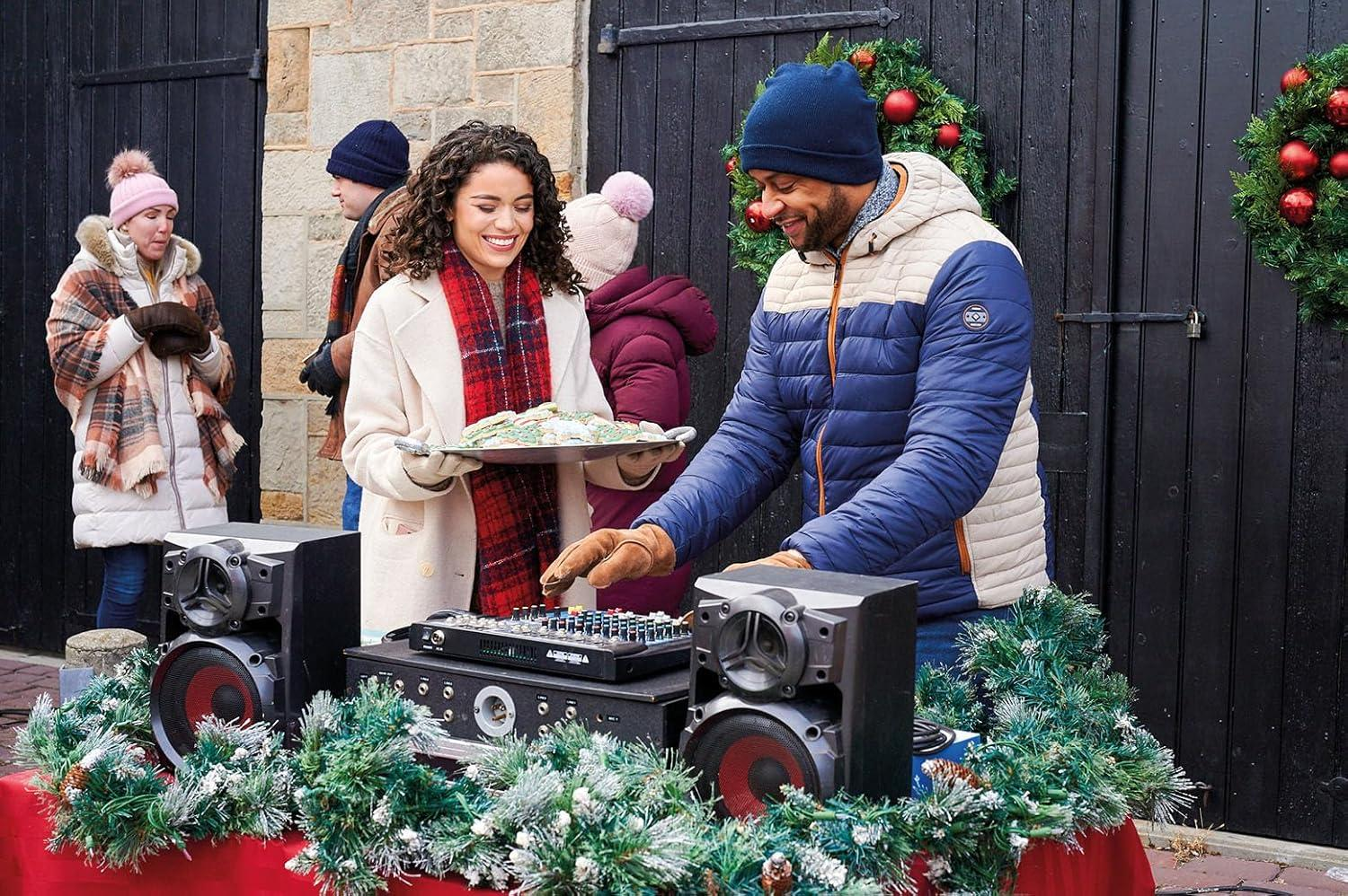 Leute in der Nähe von Lautsprechern und einem DJ-Mischpult. Eine Frau hält ein Tablett mit Keksen. Weihnachtskränze sind sichtbar. Alle tragen Winterkleidung.