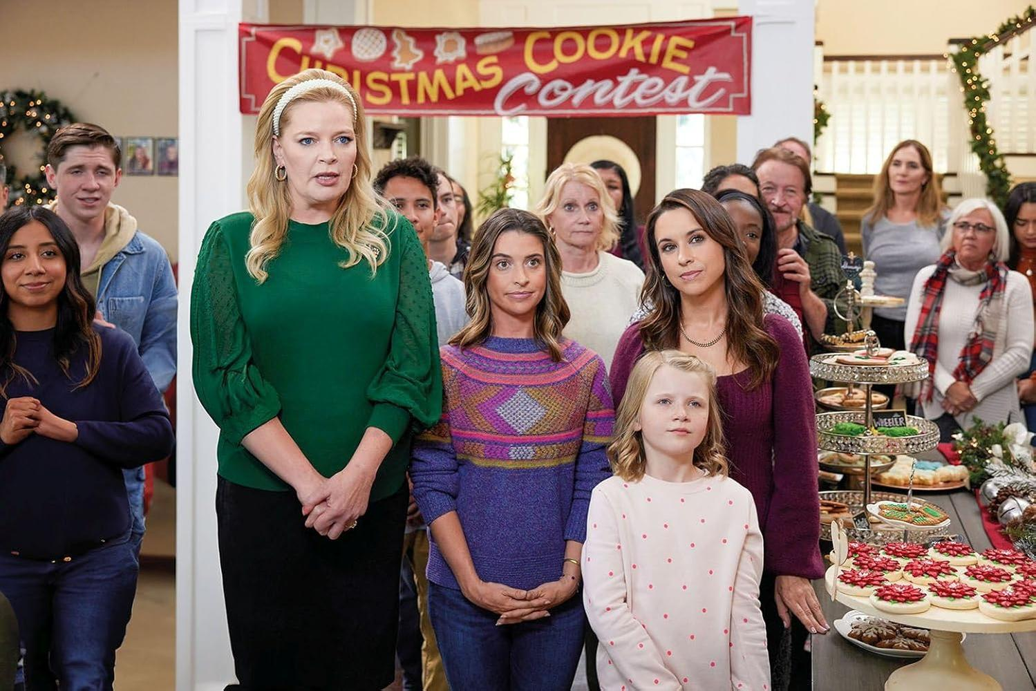 Gruppe von Menschen bei einem Weihnachtsplätzchenwettbewerb. Einige schauen in die Kamera. Kekse werden ausgestellt. Ein Banner lautet 'Christmas Cookie Contest'.