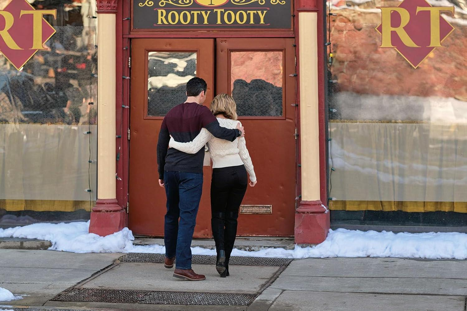 Ein Paar geht Arm in Arm auf eine Tür mit einem Schild 'Rooty Tooty' zu. Schnee säumt den Gehweg.
