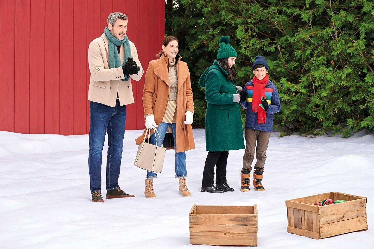 Eine vierköpfige Familie in Winterkleidung steht im Schnee in der Nähe einer roten Wand und grüner Bäume. Holzkisten sind im Vordergrund.