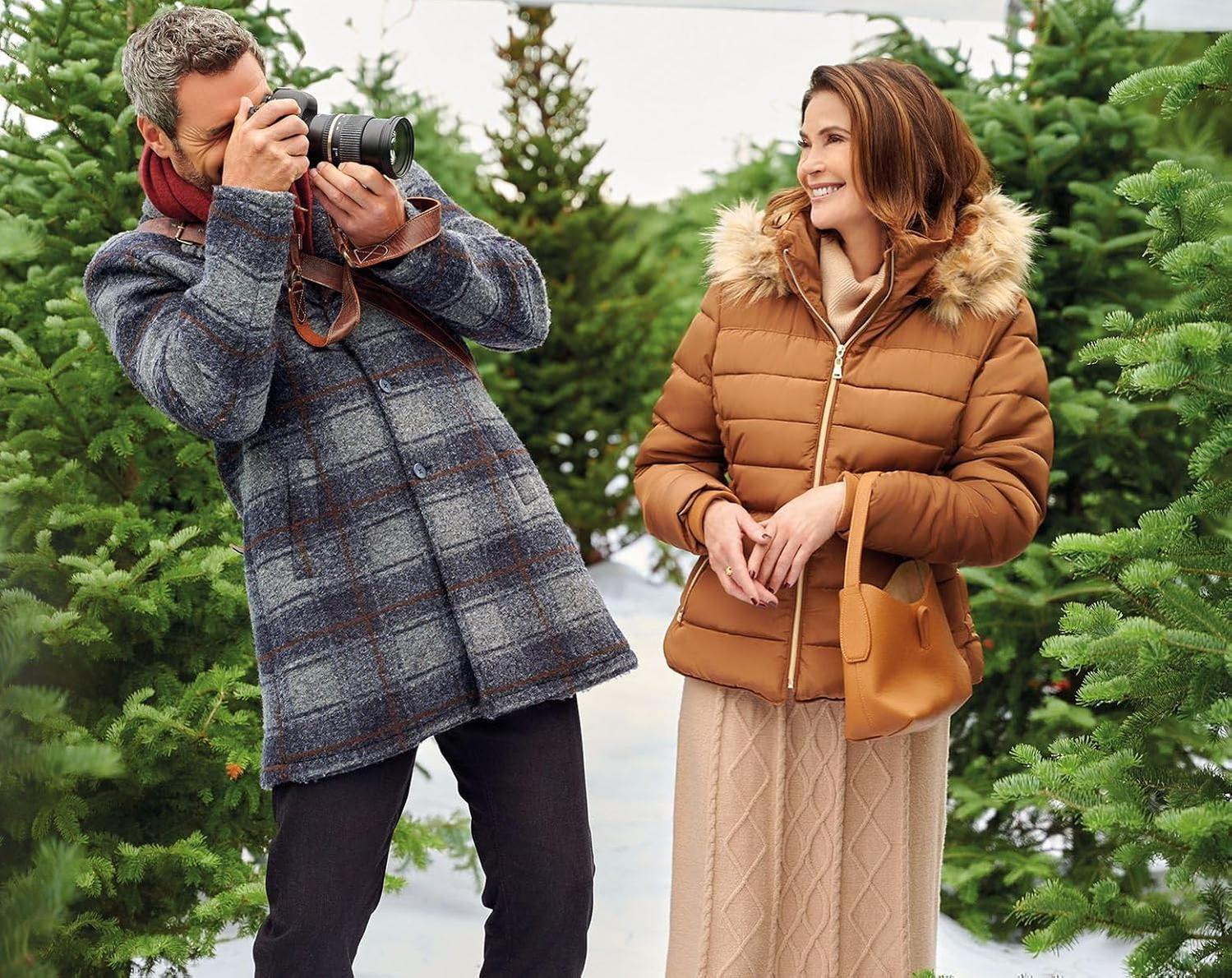Ein Mann fotografiert eine Frau. Die Frau lächelt und trägt einen braunen Mantel und eine Handtasche. Sie sind in einem schneebedeckten Gebiet.