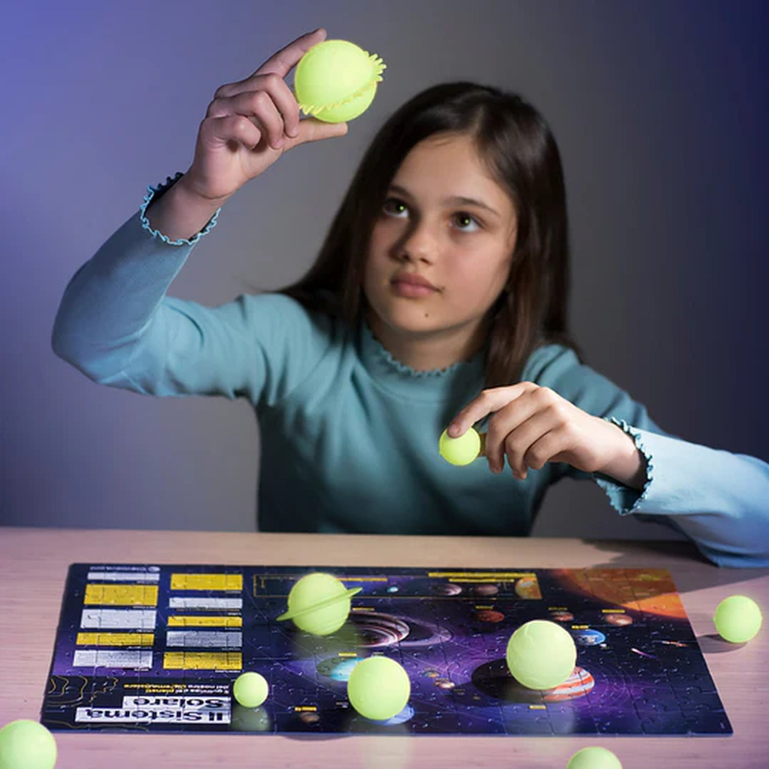 Una ragazza tiene delle sfere gialle sopra un puzzle del sistema solare. Ci sono sfere sul tavolo, alcune nelle sue mani. La scena è al chiuso.