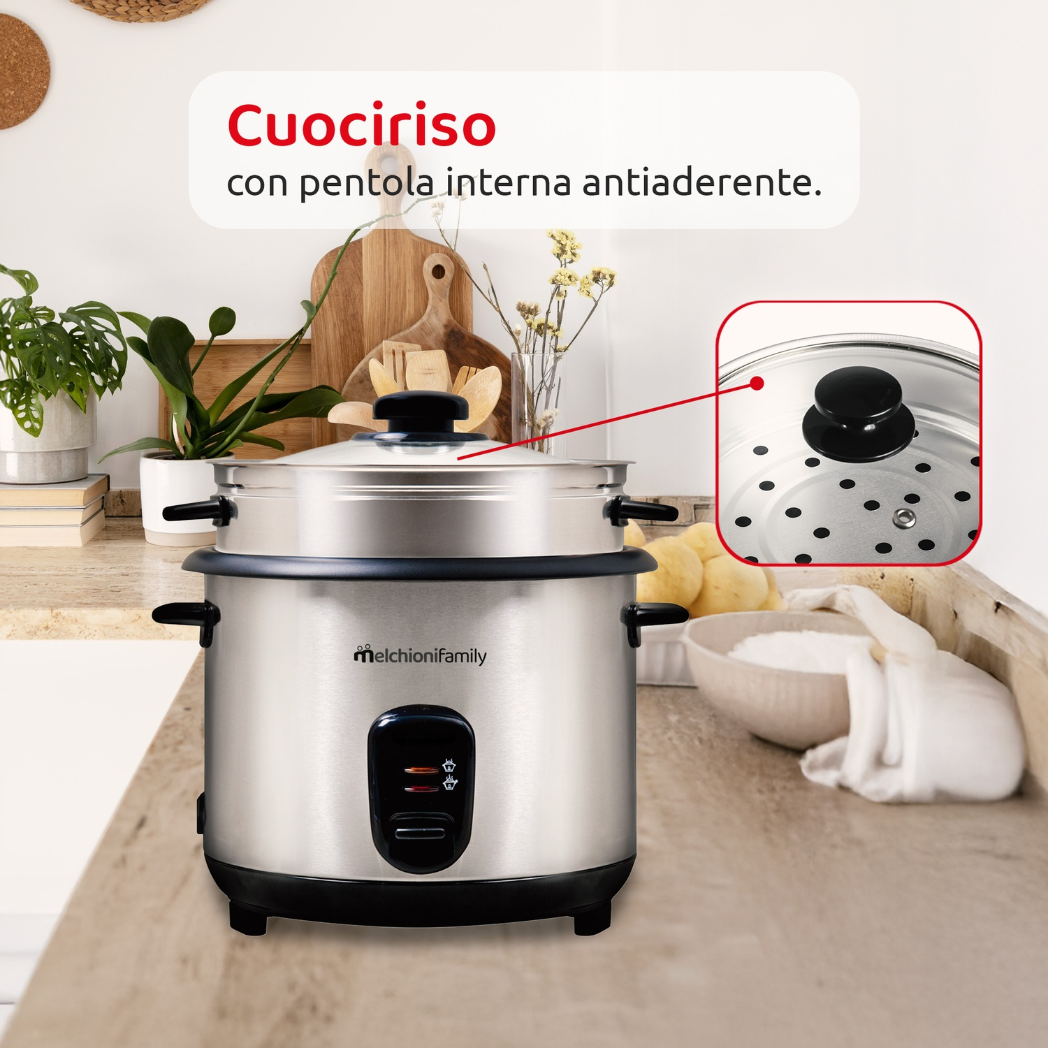 Una cuociriso argentata con manico e pannello di controllo neri è appoggiata su un piano cucina. Lo sfondo comprende piante, taglieri e ingredienti.