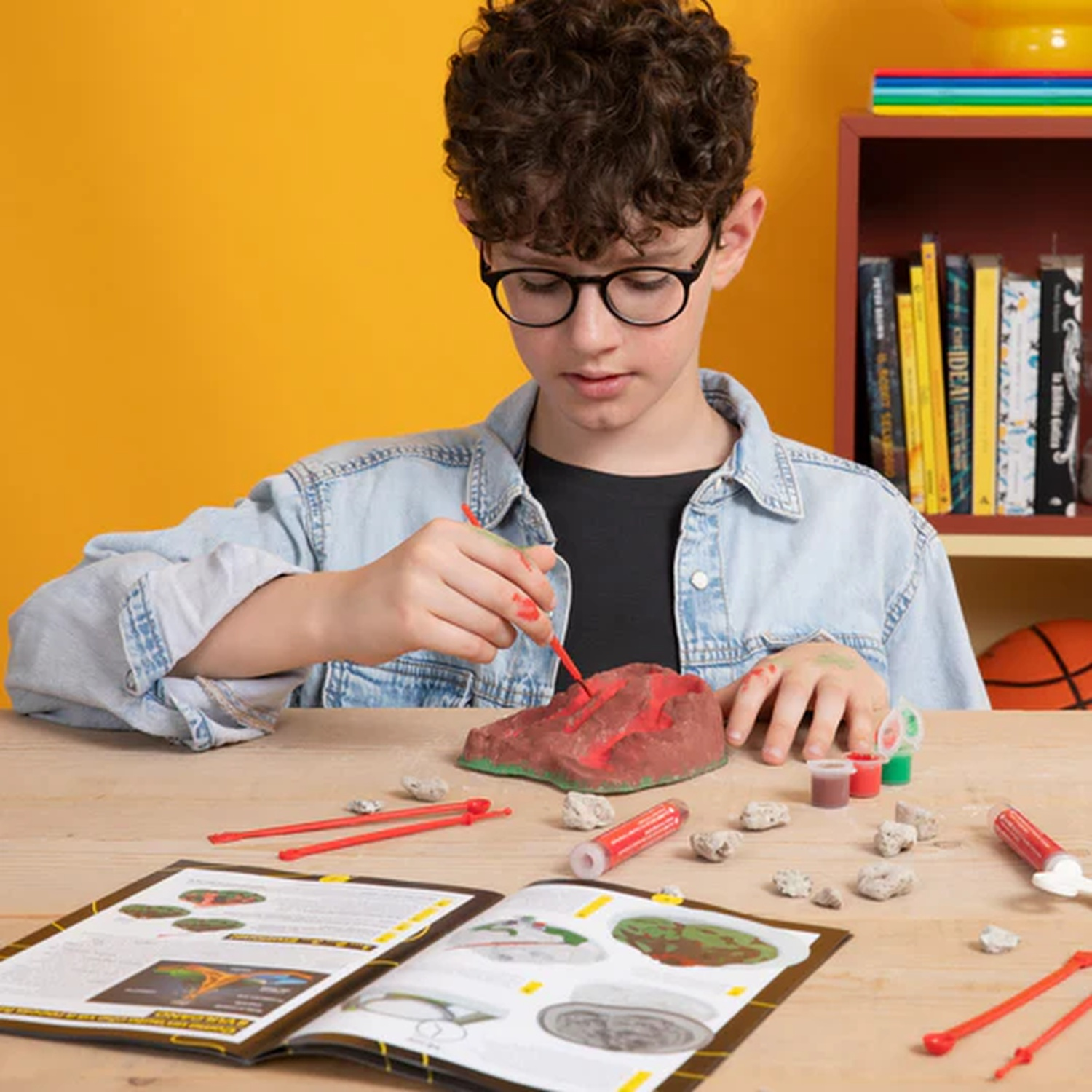 Un ragazzo con gli occhiali dipinge un modello di vulcano. Vernici, rocce e istruzioni sul tavolo. Libreria e palla sullo sfondo.