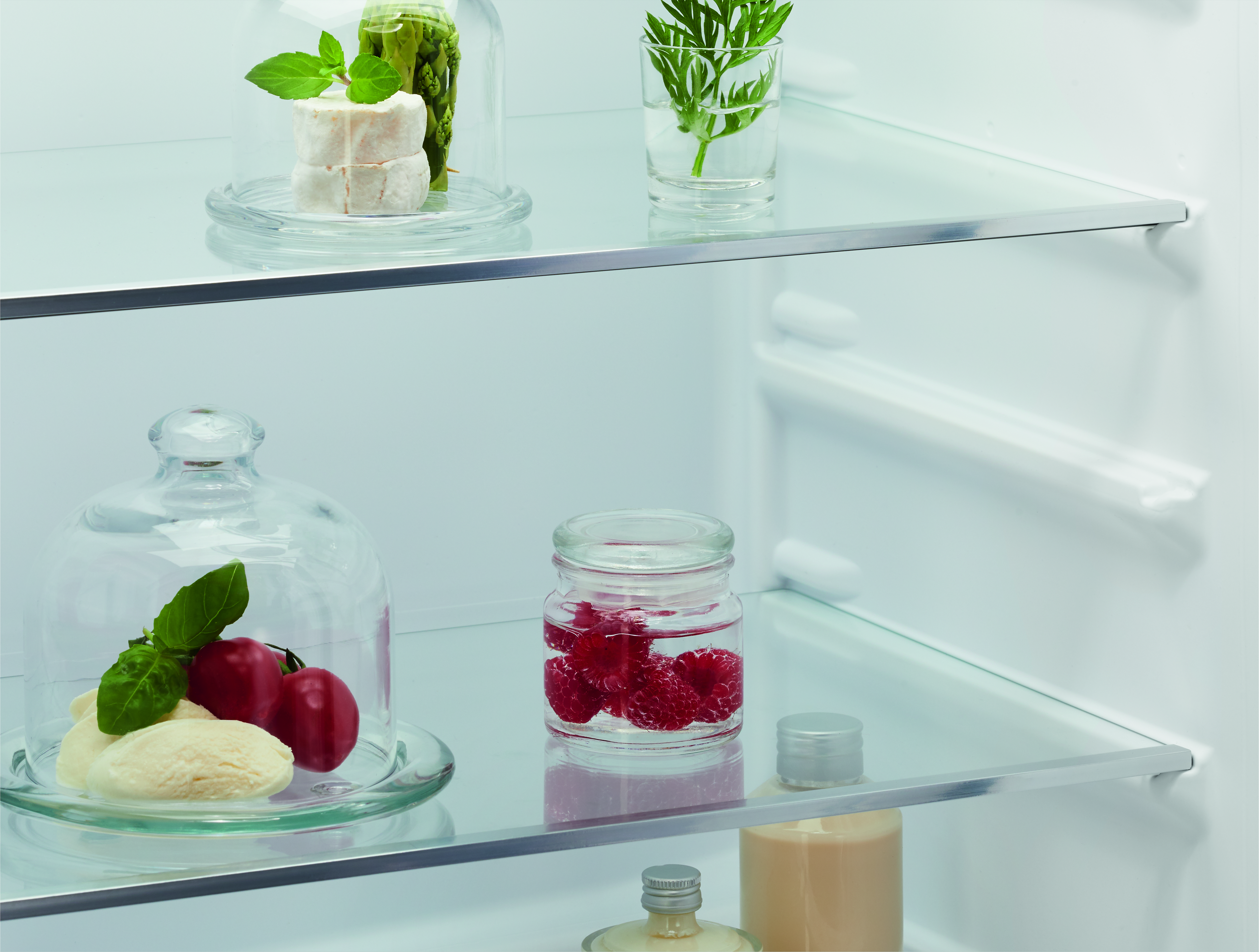 Intérieur d'un réfrigérateur avec étagères en verre. Contient des cloches en verre avec des aliments comme du fromage, des tomates et des framboises. Autres articles: asperges.