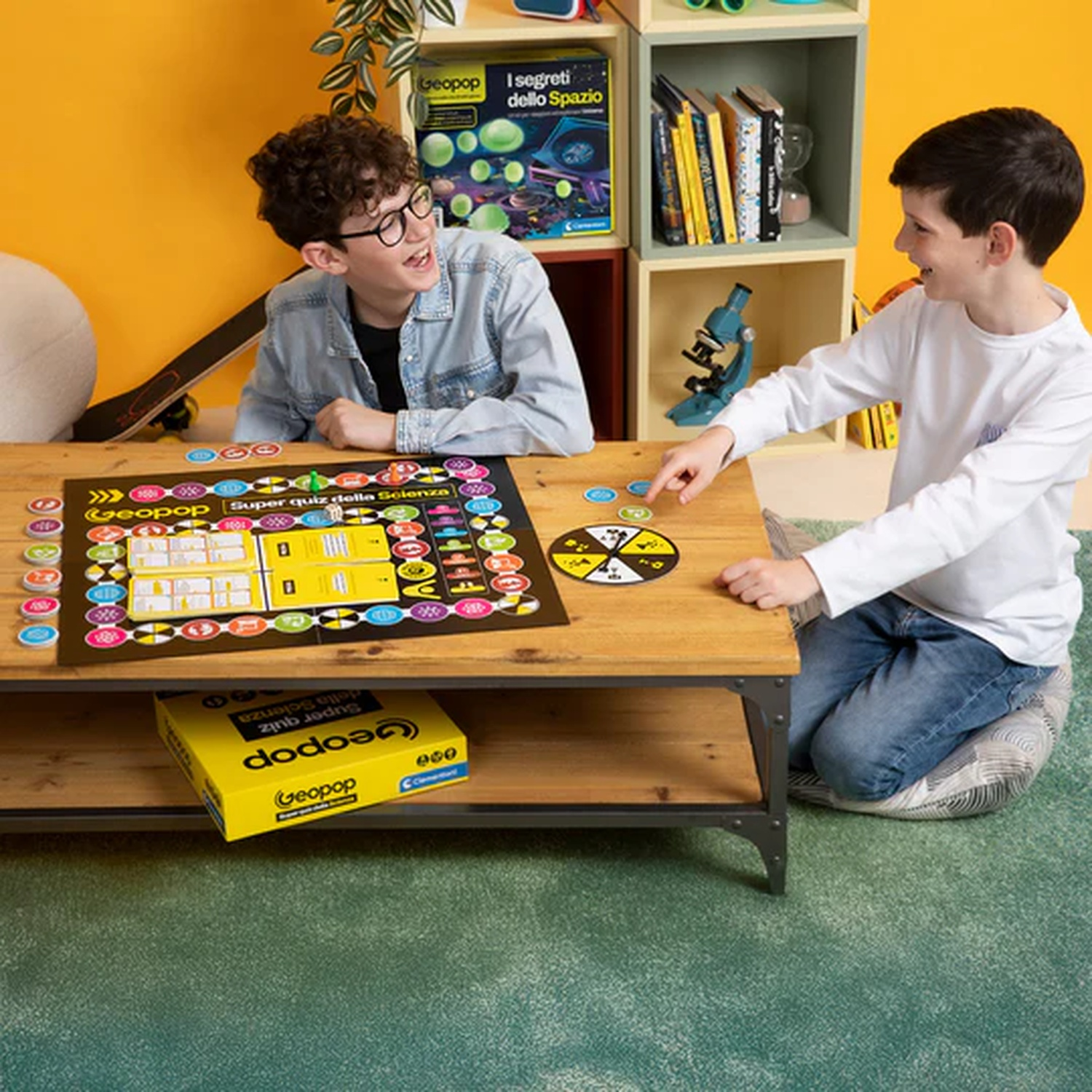 Due ragazzi giocano a un gioco da tavolo su un tavolo di legno. Un ragazzo indossa gli occhiali. La scatola del gioco è gialla. Librerie e giocattoli sullo sfondo.
