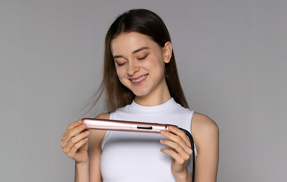 Kobieta trzyma różowo-złotą prostownicę do włosów, uśmiechając się. Ma na sobie biały top. Szare tło.