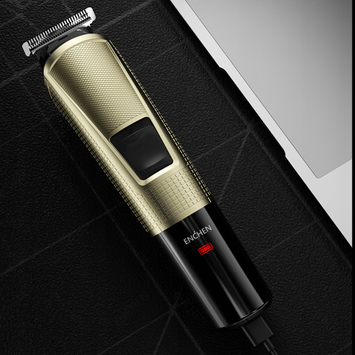 Złoto-czarna elektryczna maszynka do włosów. Trymer umieszczony jest na teksturowanej czarnej powierzchni, z laptopem w tle.