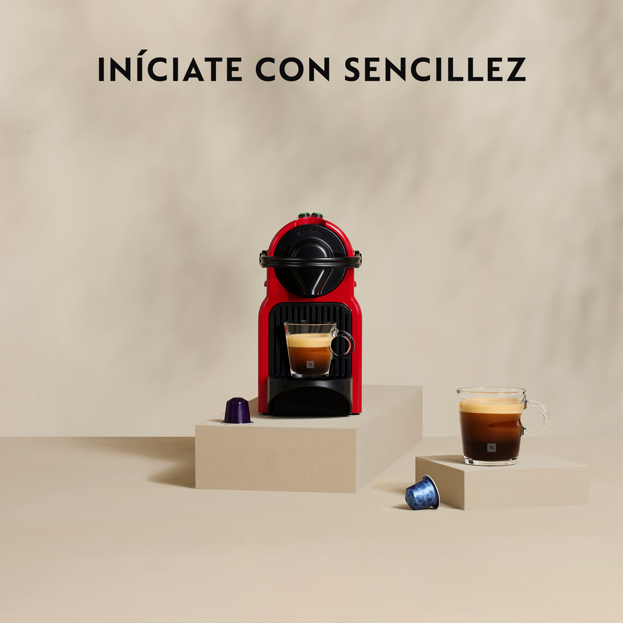 Una máquina Nespresso roja con tazas de café y cápsulas sobre una superficie beige. El texto 'INÍCIATE CON SENCILLZ' está arriba.