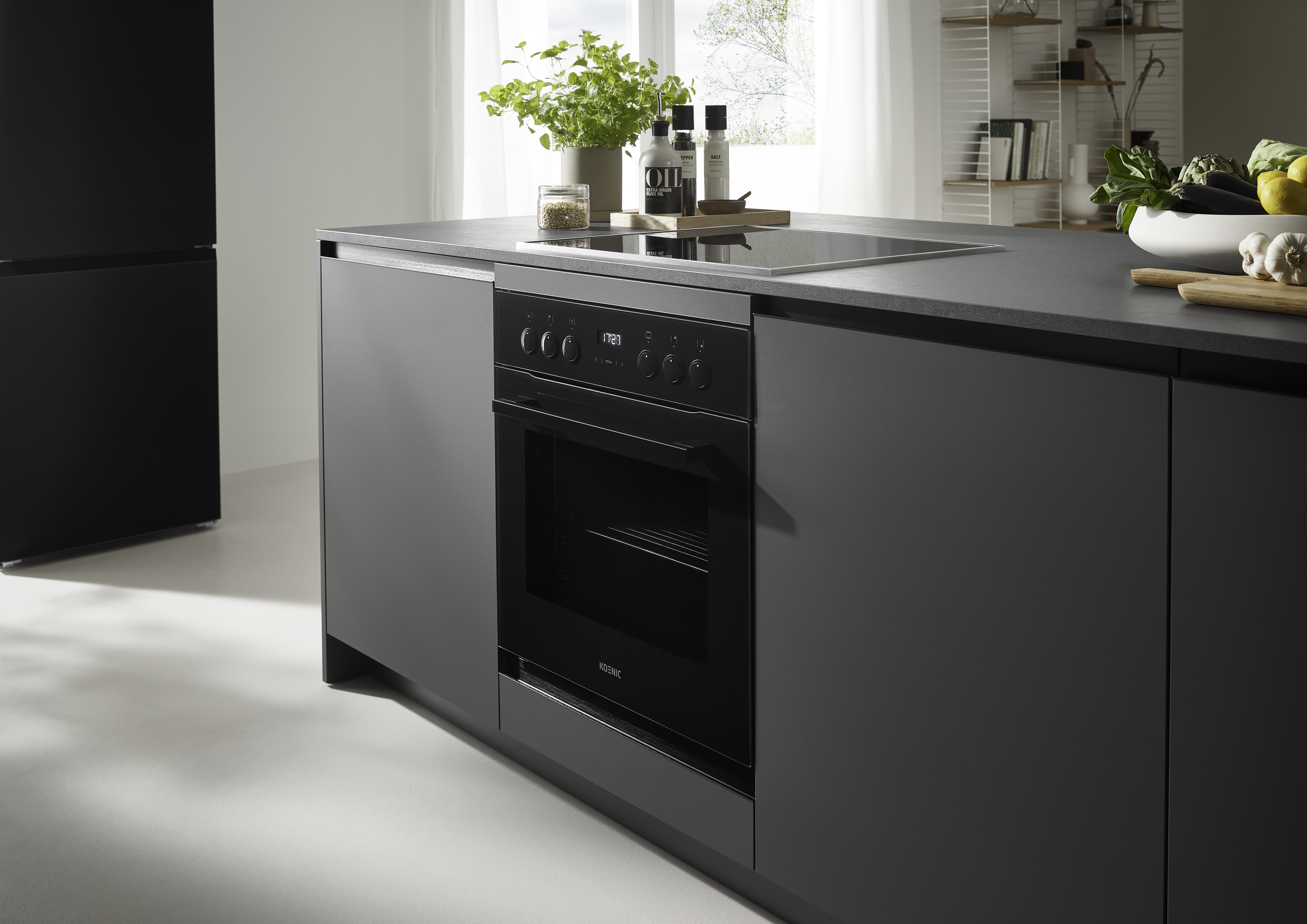 Moderne Küche mit dunkelgrauen Schränken, Backofen und Arbeitsplatte mit Pflanzen und Kochutensilien. Natürliches Licht.