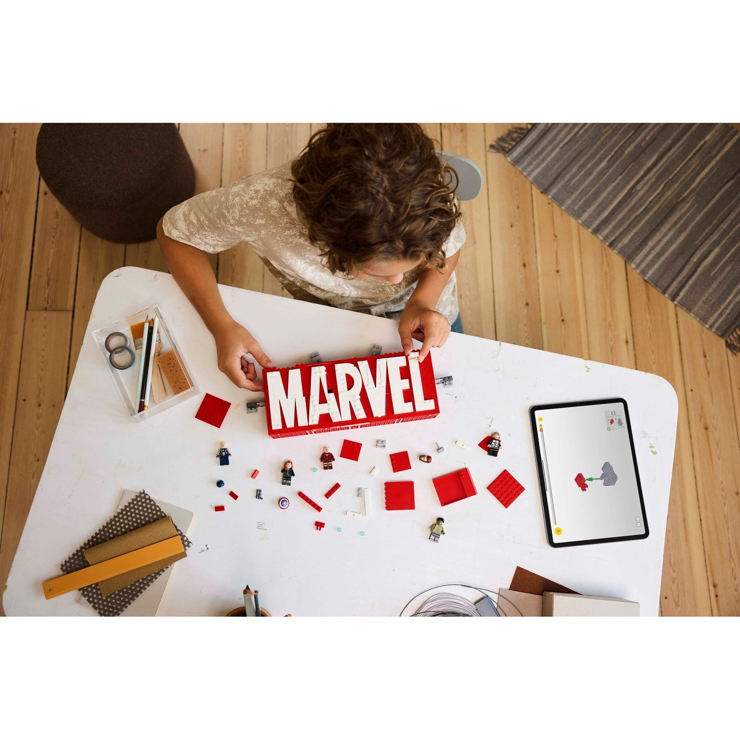 Un bambino assembla un set Lego Marvel su un tavolo bianco. Il set include mattoncini rossi, minifigure e un tablet con le istruzioni.