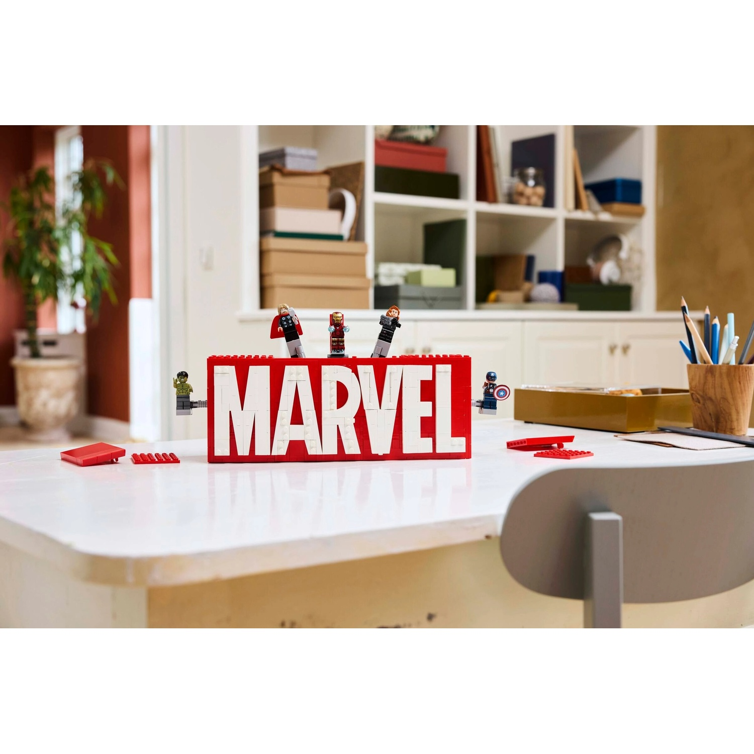 Un tavolo bianco con un cartello MARVEL rosso e personaggi Lego. Lo sfondo mostra una stanza con scaffali, una pianta e una sedia.