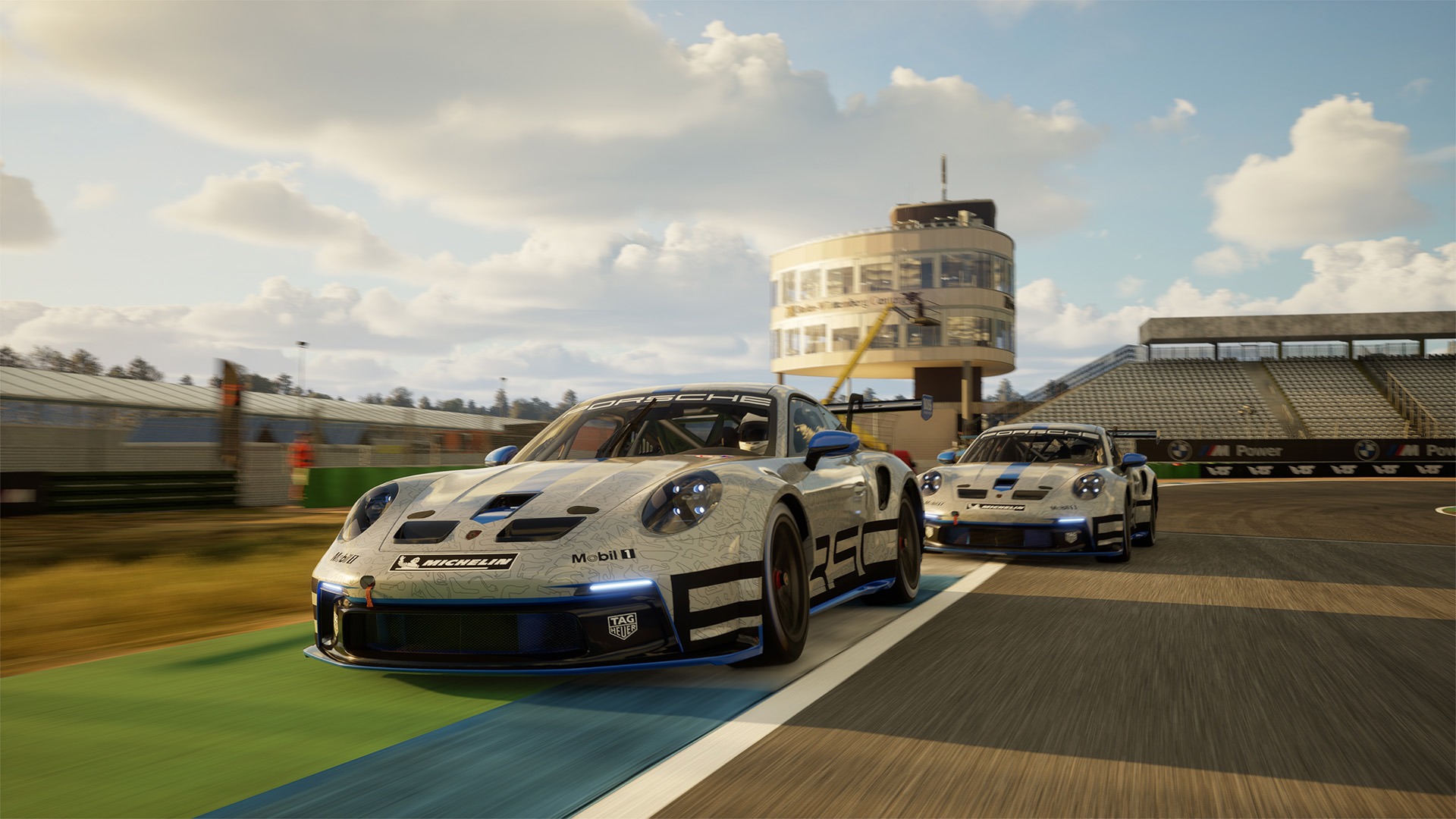 Zwei Porsche-Rennwagen auf einer Rennstrecke, einer vorne. Gebäude und Tribünen im Hintergrund, blauer Himmel mit Wolken.