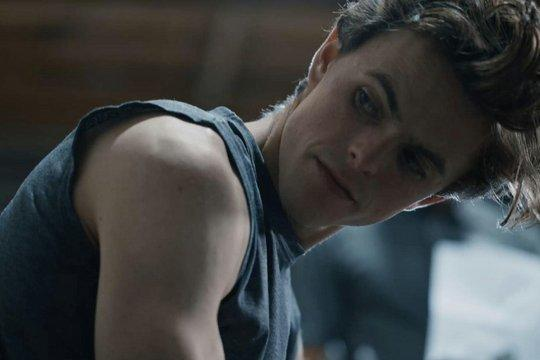 Ein junger Mann in einem grauen Tanktop blickt leicht lächelnd nach unten. Seine dunklen Haare sind gestylt, der Hintergrund ist verschwommen und dunkel.