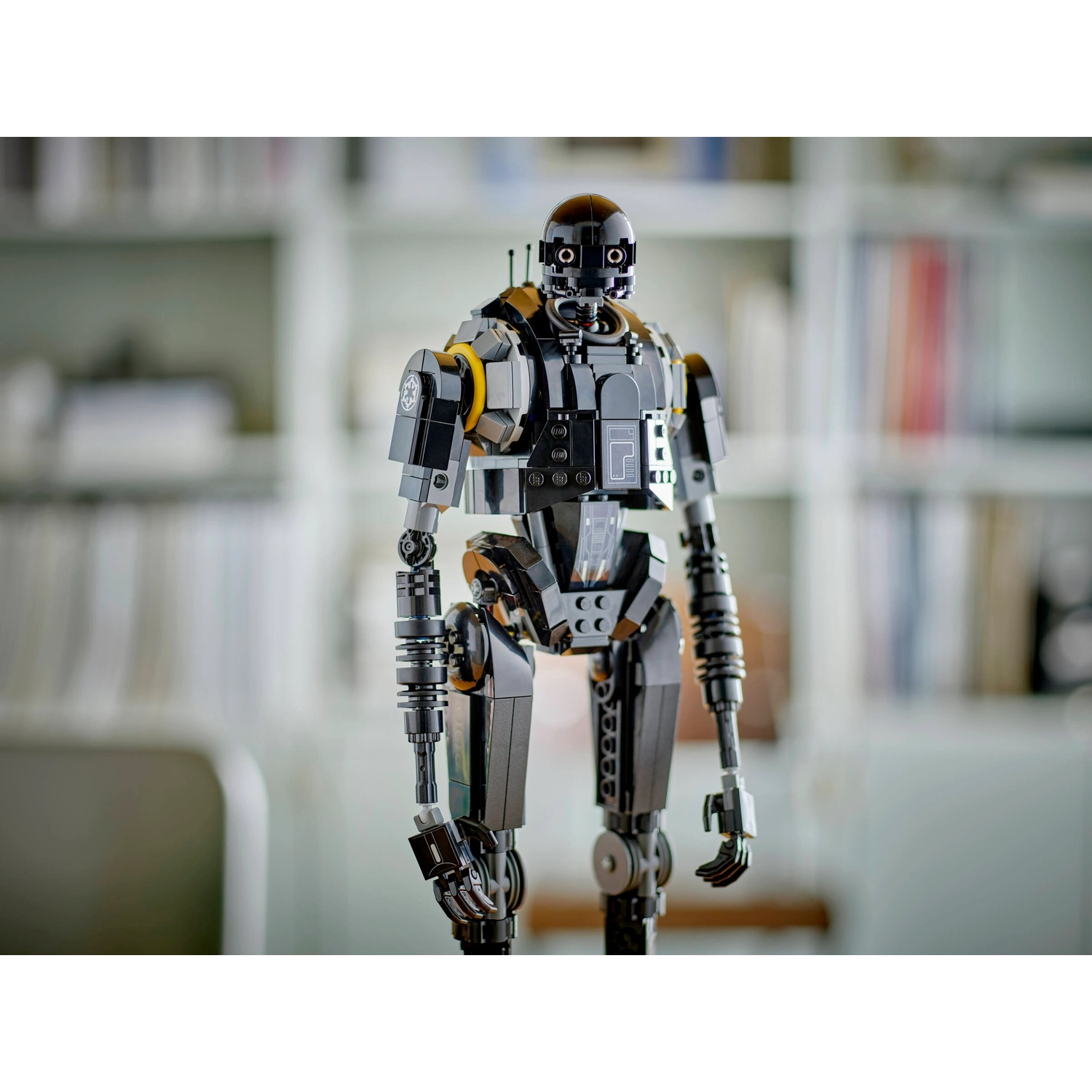 Un robot K-2SO Lego nero è in piedi, dettagliato con accenti grigi e gialli. Lo sfondo è una libreria sfocata.