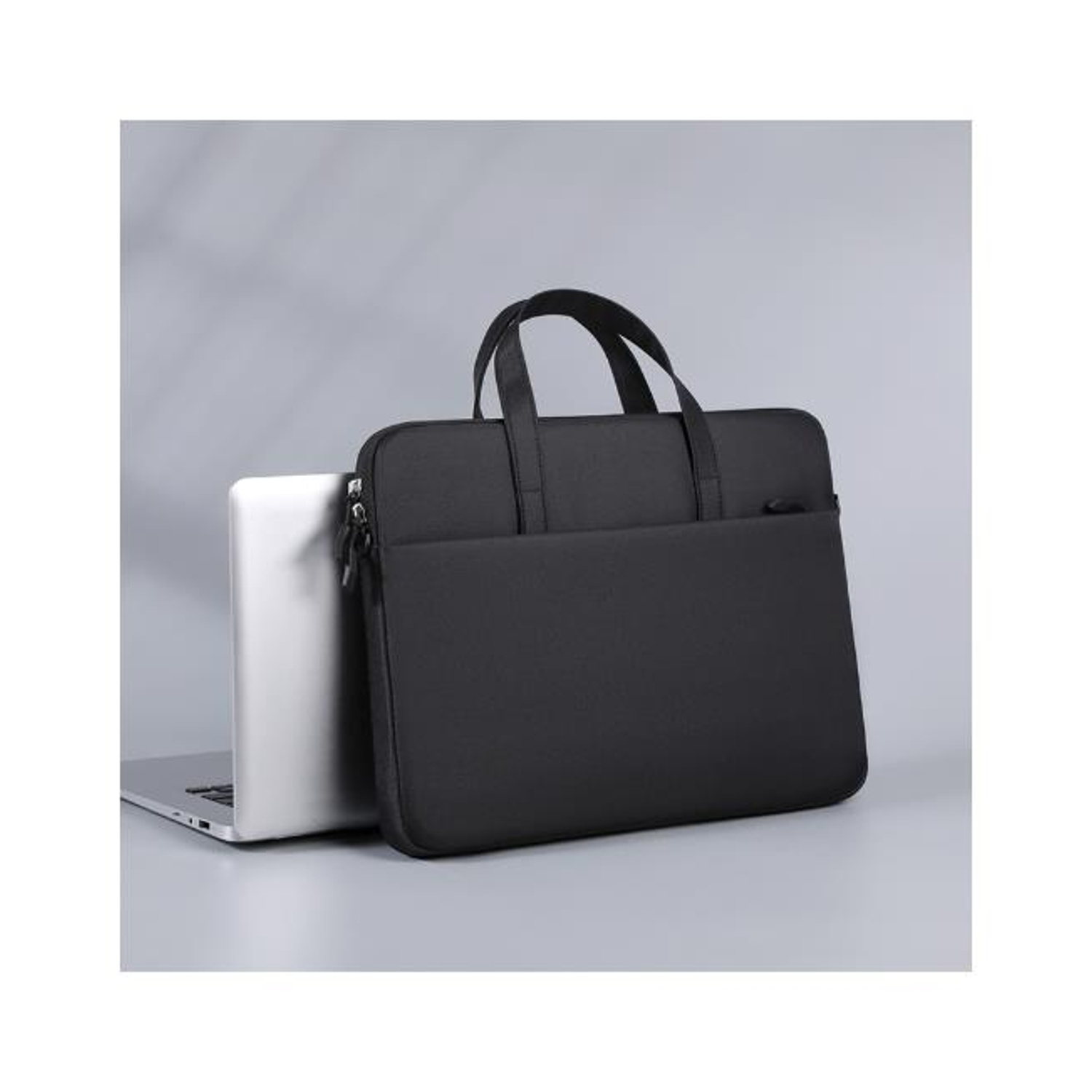Una borsa nera per laptop accanto a un laptop argentato. Lo sfondo è grigio chiaro, creando un'estetica moderna e pulita.