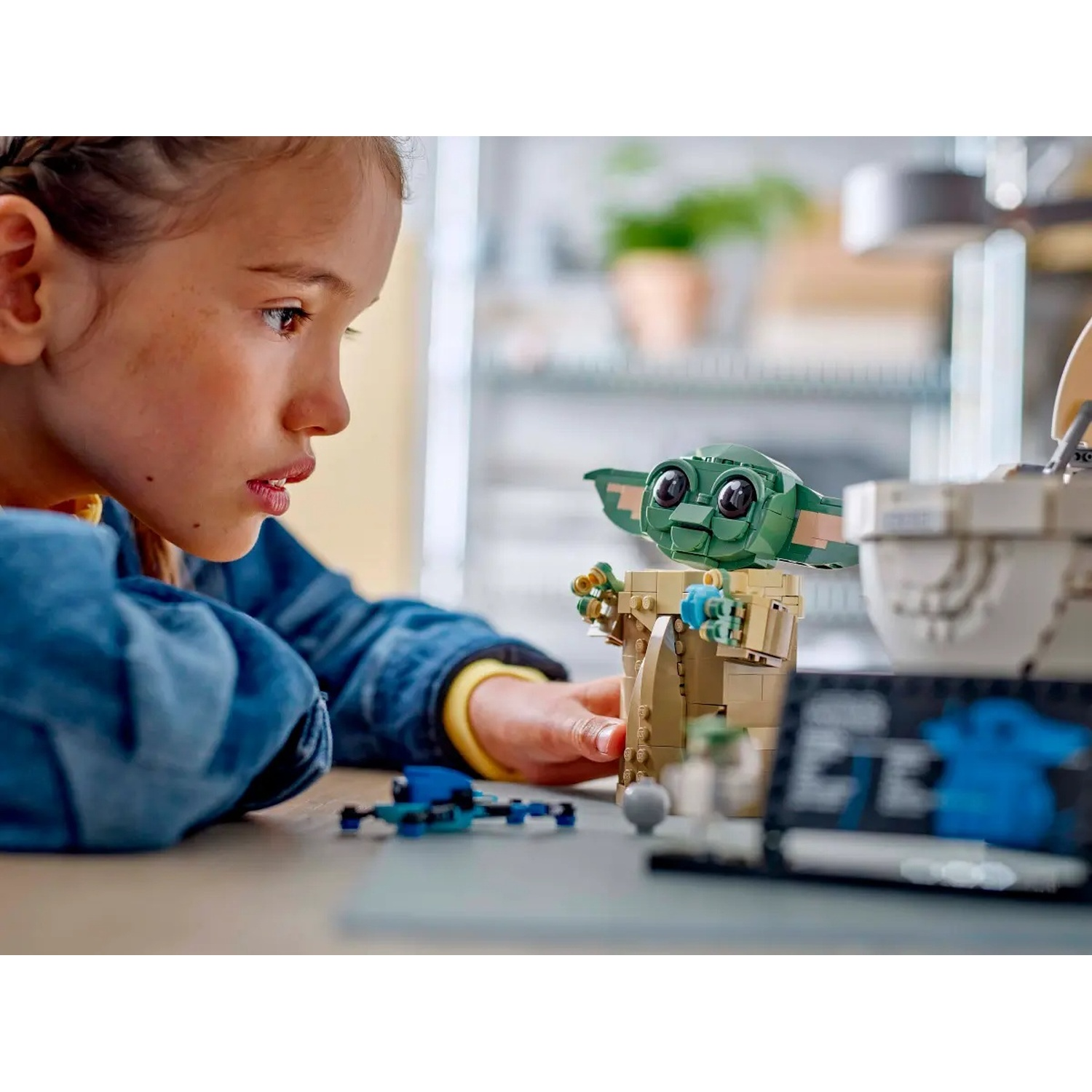 Una ragazza guarda un Baby Yoda Lego. La scena comprende una nave Lego blu, un Baby Yoda Lego e una parte di una nave Lego.