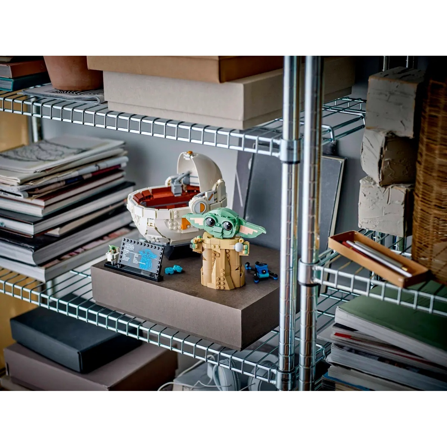 Un Baby Yoda Lego è in piedi su uno scaffale con la sua culla. Libri e scatole sullo sfondo. Un cartello e altri pezzi Lego sono visibili.
