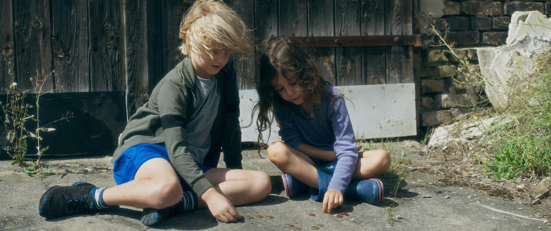 Zwei Kinder sitzen und zählen Münzen auf Beton. Einer hat blondes Haar und eine grüne Jacke. Der andere hat dunkles Haar und ein blaues Hemd.
