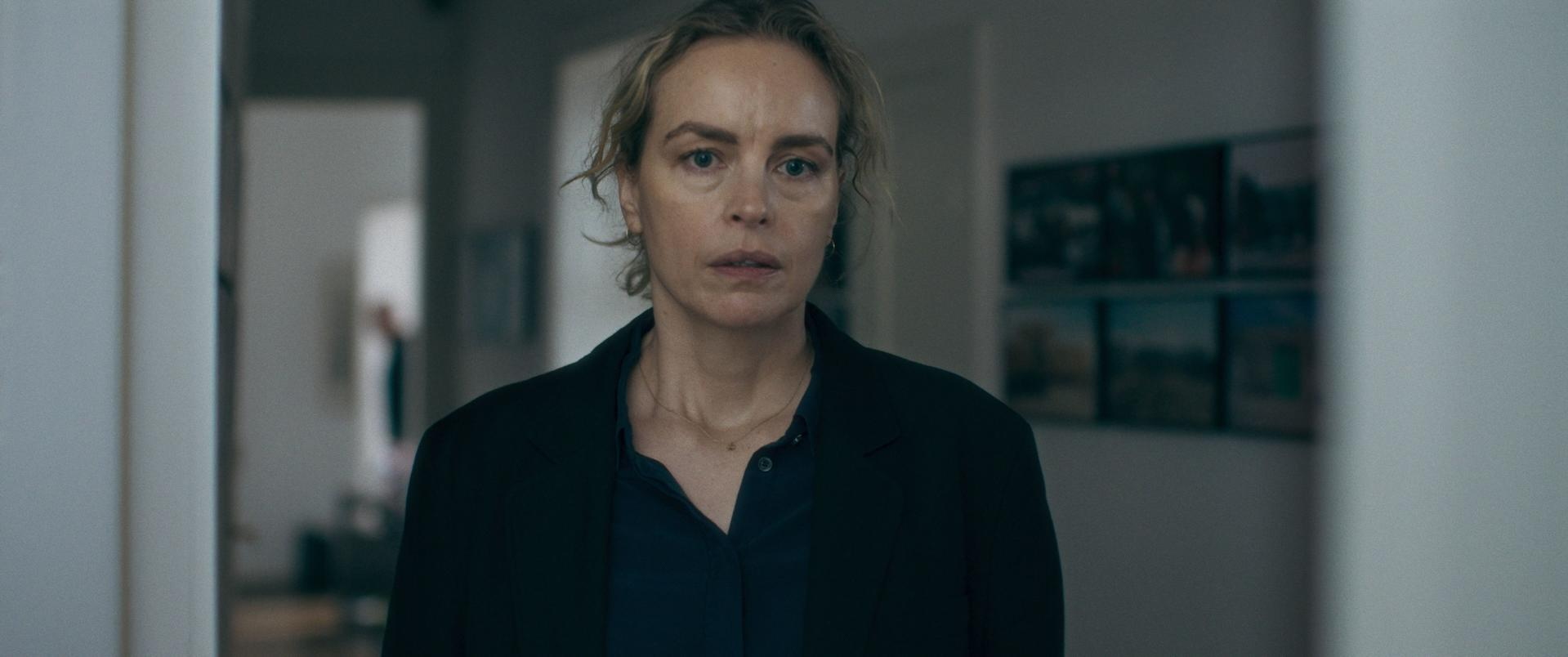 Eine Frau mit blonden Haaren und einem schwarzen Blazer blickt direkt in die Kamera. Sie steht in einem Flur.