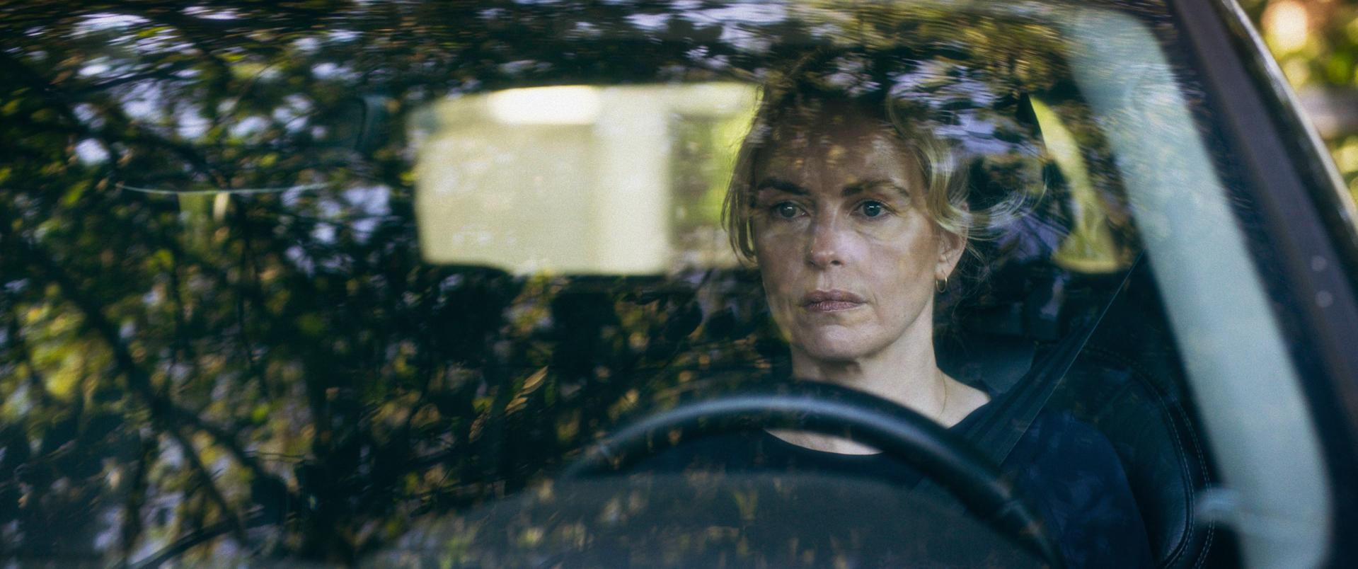 Eine Frau fährt Auto, durch die Windschutzscheibe gesehen. Sie blickt direkt in die Kamera. Bäume spiegeln sich.