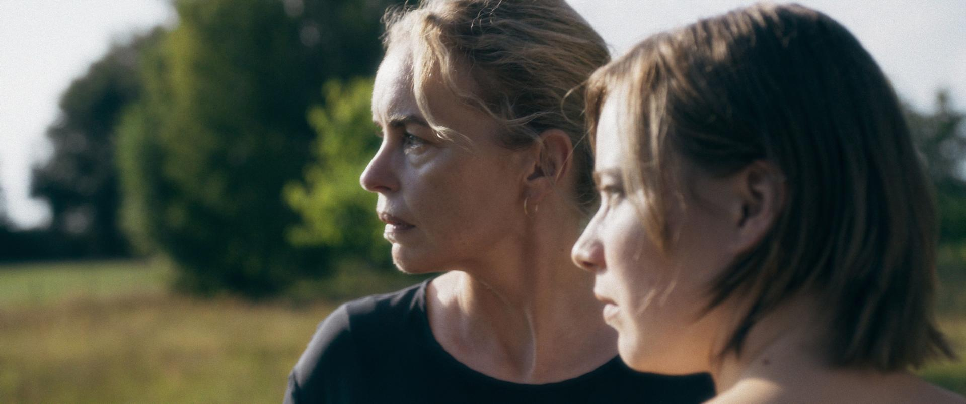 Zwei Frauen im Profil im Freien. Eine hat blondes Haar und ein schwarzes Hemd. Die andere hat braune Haare. Grüne Bäume im Hintergrund.