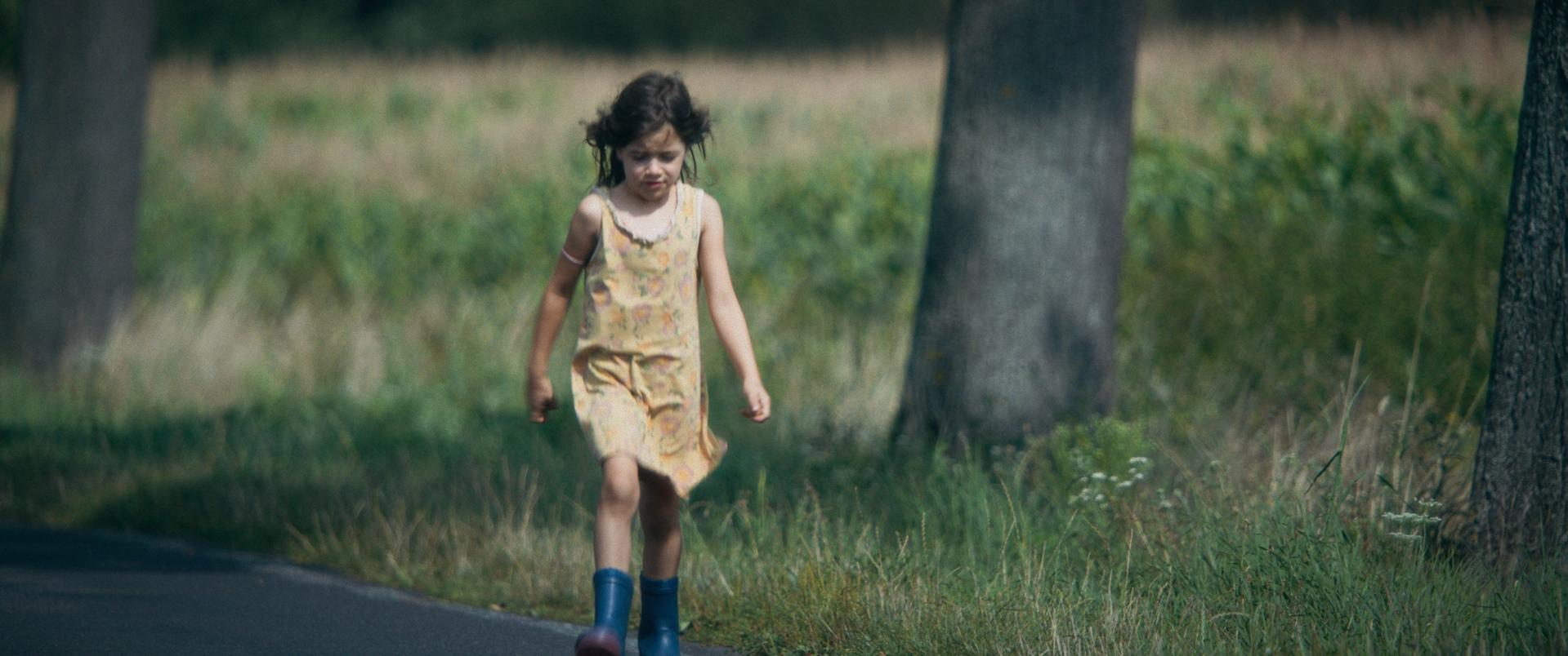 Ein junges Mädchen geht auf einer Straße. Sie trägt ein gelbes Kleid und blaue Stiefel. Bäume und grünes Gras sind im Hintergrund.