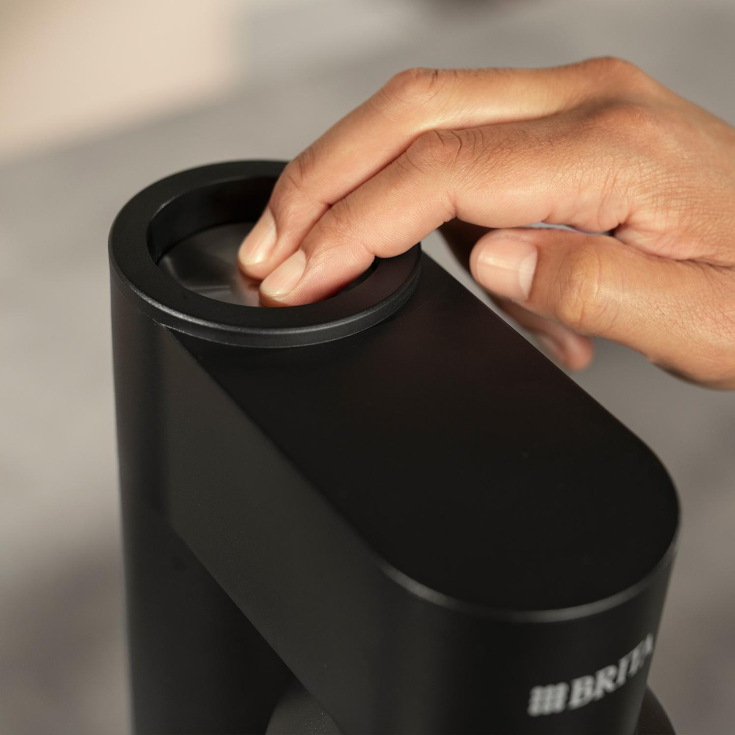 Ręka naciska palcem na czarny dozownik wody. Dozownik ma zakrzywioną górę i widoczne jest logo Brita.